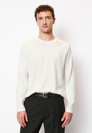 Long-sleeve, crew-neck shirt in white cotton, featuring raglan sleeves and a relaxed fit, paired with black trousers and a black belt.