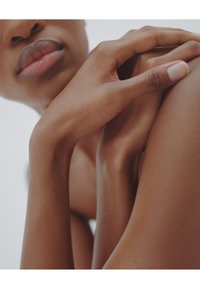 Gros plan sur des tons de peau lisses ; des mains reposent doucement sur une épaule. Texture subtile et contours naturels mis en valeur sur un fond clair.