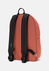 Mochila de tela color coral naranja, con correas de hombro acolchadas negras, un asa superior y un bolsillo lateral. Textura suave con detalles del logo.