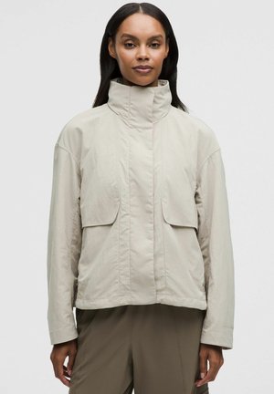 Femme portant une veste beige clair à col montant avec poches à rabat et manches longues, debout devant un fond uni.