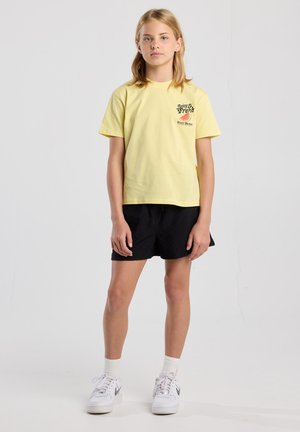 Yellow cotton t-shirt featuring a graphic print on the left chest, paired with black shorts and white sneakers. Simple, casual design.