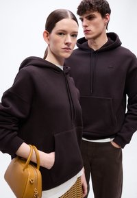 Νεαρή γυναίκα και άνδρας φορούν σκούρες κουκούλες, η γυναίκα κρατά κίτρινη τσάντα, στέκονται μπροστά σε απλό φωτεινό φόντο.