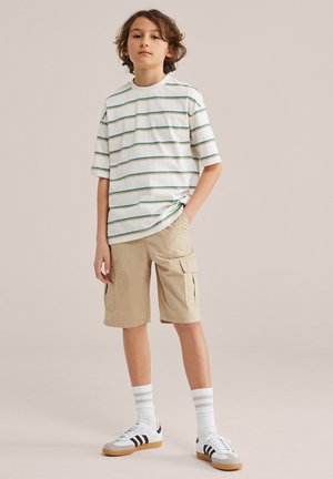 Garçon debout les mains dans les poches, portant un t-shirt blanc rayé, un short cargo beige, des chaussettes blanches et des baskets blanches à rayures noires.