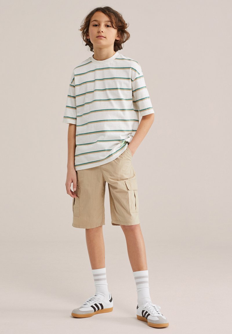 Junge steht mit Händen in den Taschen, trägt ein gestreiftes weißes T-Shirt, beige Cargo-Shorts, weiße Socken und weiße Turnschuhe mit schwarzen Streifen.