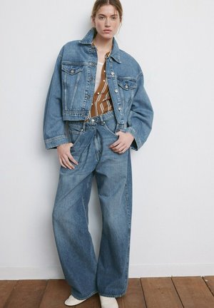 Mladá žena v nadmernej modrej rifľovej bunde a širokých džínsoch, doplnená o pásikavú hnedo-bielu košeľu a biele čižmy.