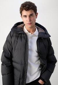 Schwarze Steppjacke mit Kapuze, ausgestattet mit einer gesteppten Textur, einem Frontreißverschluss und einer Brusttasche. Getragen über einem weißen Poloshirt.