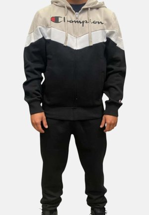 Persona che indossa una felpa Champion a blocchi di colore beige, bianco e nero con pantaloni neri abbinati e scarpe nere, in piedi con le mani rilassate.