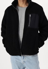 Schwarze Fleecejacke mit durchgehendem Reißverschluss und einer Brusttasche, aus strukturiertem Material und einem kontrastierenden matten Panel auf der Tasche.