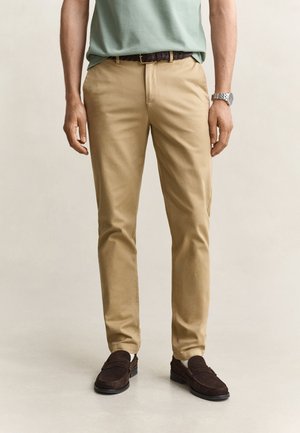Mann trägt beige Chinos, dunkelbraunen geflochtenen Gürtel, braune Wildleder-Loafer und eine silberne Uhr, steht vor einem einfarbigen hellen Hintergrund.