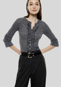 Maglia grigia a maniche lunghe con placca arricciata e chiusura con bottoni, abbinata a pantaloni a vita alta con sottili macchie, e cintura nera.