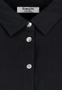 Black collared shirt made of textured fabric, featuring five buttons with a pearl-like finish and a label reading "Sublevel," size M/L.