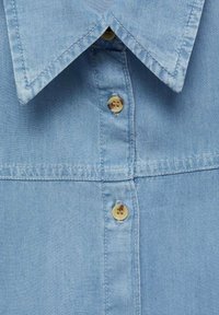 Light blue denim shirt with visible collar, horizontal seam, and three beige buttons along the front placket.