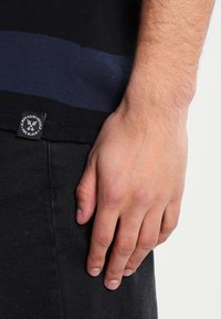 Sort T-shirt med en blå vandret stribe, der har en vævet etiket nær kanten. Hånd hviler på stoffet, hvilket viser tekstur og pasform.