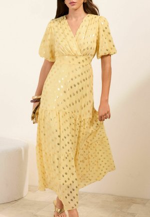 Frau trägt ein gelbes Wickelkleid mit Puffärmeln und metallisch goldenen Punkten, dazu Armreifen und goldene Sandalen.