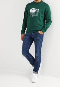 Grön sweatshirt med vitt grafiskt mönster, kombinerad med blå jeans och vita sneakers. Tyget verkar mjukt och avslappnat.