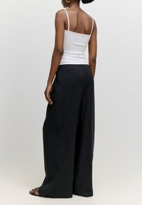 Zwarte wijde broek met een gladde textuur, gecombineerd met een strak wit spaghetti-topje. Model gezien vanachter.