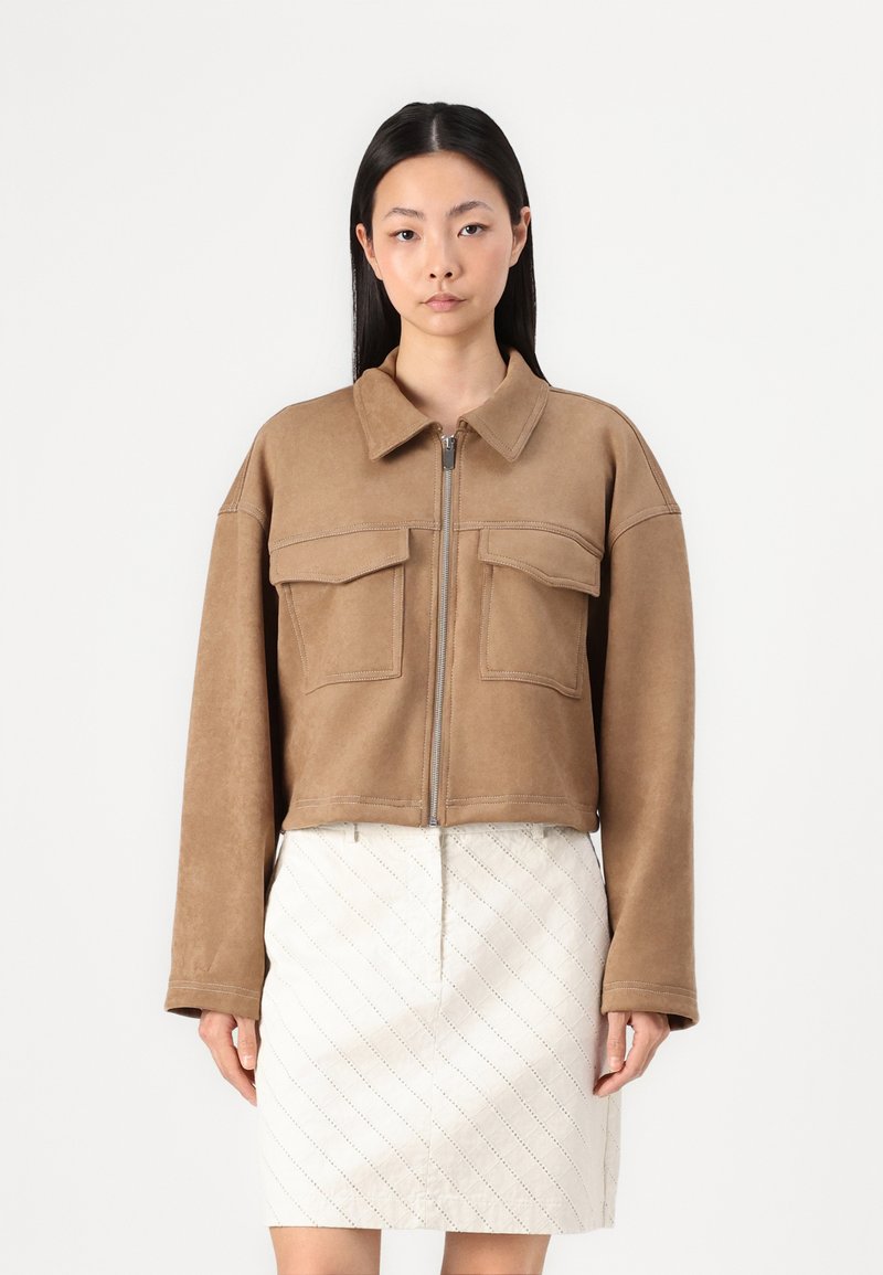 Bruine cropped jas van zacht materiaal met een ritssluiting aan de voorkant, grote borstzakken en lange mouwen, gecombineerd met een witte rok met diamantpatroon.