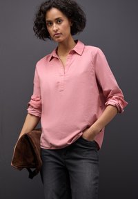 Lichtroze blouse met een kraag en knoopsluiting, opgerolde mouwen, gestructureerde stof. Draagt een donkergrijze jeans. Bruine jas in de hand.