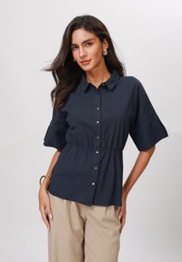 Blusa blu navy con bottoni frontali, colletto, vita elastica e maniche corte; texture liscia con leggero lucido. Pantaloni beige visibili.