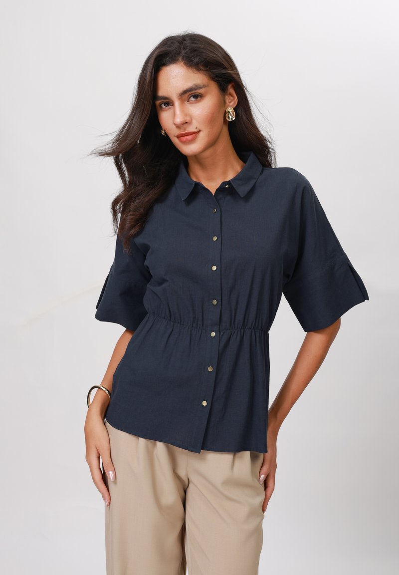 Blusa blu navy con bottoni frontali, colletto, vita elastica e maniche corte; texture liscia con leggero lucido. Pantaloni beige visibili.