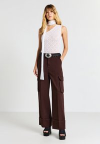 Giovane donna che indossa un top bianco in pizzo senza maniche con sciarpa abbinata, pantaloni cargo brown a gamba larga con cintura nera e tacchi a piattaforma.