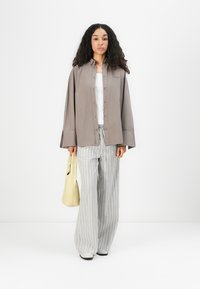 Gray oversized button-up shirt, white tank top, gray and white striped wide-leg pants, holding a pale yellow bag, standing on a white background.