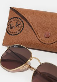Occhiali da sole Ray-Ban con lenti color marrone, montatura in metallo dorato e un astuccio in pelle marrone textured con logo stampato e chiusura a bottone.
