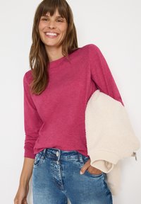 Långärmad fuchsia-topp i lätt tyg, tillsammans med ljusblå denimjeans. Modellen håller en krämfärgad, texturerad jacka.