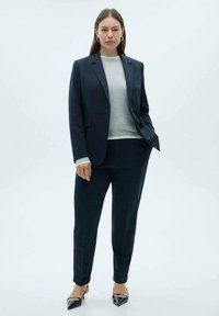 Traje de corte ajustado en azul marino con una chaqueta estructurada y pantalones tapered a juego. Llevado sobre un suéter gris claro, con tacones negros puntiagudos.
