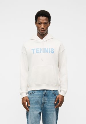 Giovane uomo con felpa bianca con cappuccio con la scritta "TENNIS" in lettere azzurro chiaro, abbinata a jeans di denim azzurro chiaro, in piedi su sfondo bianco.