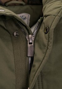Olivegrüne Jacke mit einem Metallreißverschluss, Druckknopfverschlüssen und strukturiertem Stoff. Verfügt über ein gesteppte Innenfutter und ein Etikett im Kragen.