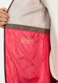 Fleece-gevoerde jasje met een beige buitenlaag en rode voering. Toont waterafstotende, winddichte en dons-vrije symbolen nabij de rits.