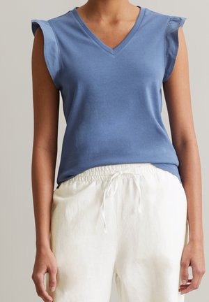 Jasnoniebieski bawełniany top bez rękawów z dekoltem w serek i falbankami na ramionach, w połączeniu z luźnymi białymi lnianymi spodniami.