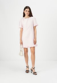 Vestido rosa claro com mangas bufantes curtas e uma bainha com babados. A modelo usa sandálias de salto pretas com bolinhas, segurando uma pequena bolsa branca.