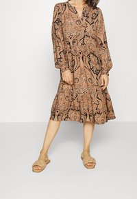 Vestido de paisley marrón y negro con mangas largas, escote en v, cintura ajustada y falda en capas. Combinado con sandalias beige de tipo slip-on.