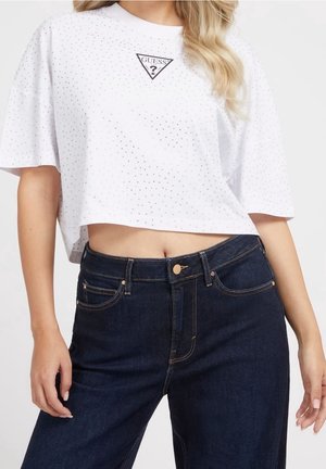 Vrouw draagt een wit cropped T-shirt met kleine stippen en een Guess-logo, gecombineerd met een high-waisted donkerblauwe spijkerbroek.