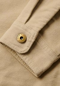 Close-up van een beige stof met een zachte textuur. Bevat een knop met de inscriptie "SUPERDRY GOODS" en een zwart oogje, bevestigd aan de mouw.