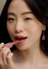 Flytende leppestift i en rosa tube påført leppene. Modell med langt mørkt hår, iført rosa bjørneformede øredobber, har et naturlig sminkelook.