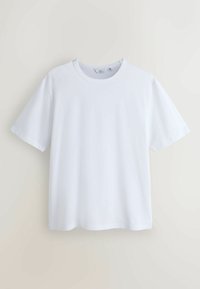 T-shirt blanc uni à manches courtes avec col rond, suspendu contre un mur beige, avec un petit logo discret sur la poitrine.