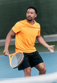 Orange athletic shirt and black shorts on a man holding a tennis racket. The background features a tennis court with green fencing.