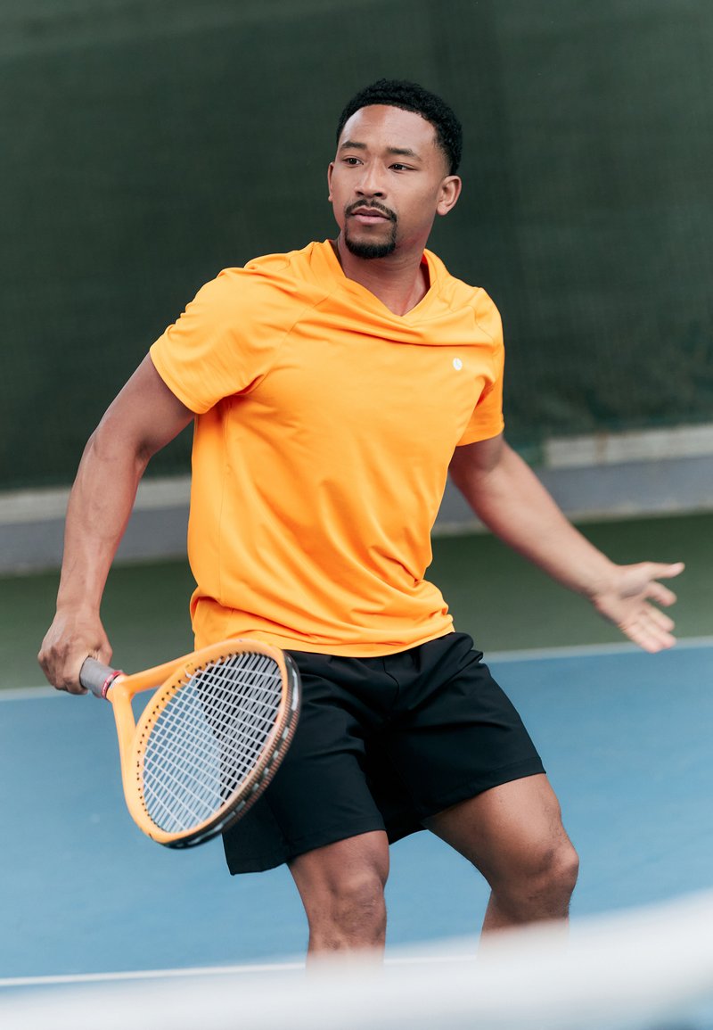 Oranges Sporthemd und schwarze Shorts an einem Mann, der einen Tennisschläger hält. Im Hintergrund ist ein Tennisplatz mit grünem Zaun zu sehen.