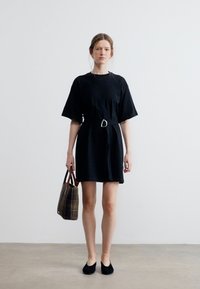 Γυναίκα που στέκεται μέσα σε εσωτερικό χώρο, φορώντας μαύρο φόρεμα με ζώνη και μαύρα παπούτσια, κρατώντας καφέ τσάντα καρό στο δεξί της χέρι.