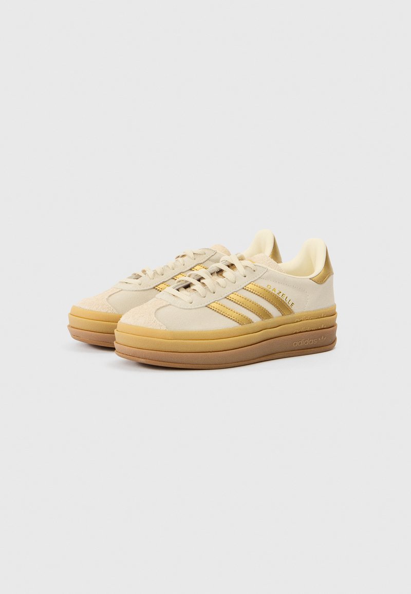 Beige Wildleder-Sneaker mit drei gelben Streifen, abgerundetem Zehenbereich, dicker Gummisohle und gepolstertem Innenfutter. Verfügt über dezente Markenkennzeichnung an der Seite.