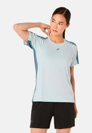 Mujer de cabello oscuro con una camiseta deportiva de manga corta azul claro y pantalones cortos negros, estirando el cuello con una mano detrás de él.