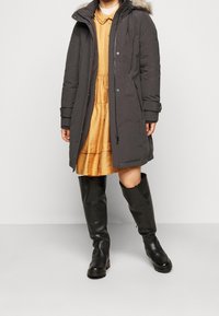 Kvinna som bär en svart knälång parkas med pälsfoder, klänning i senapsgul färg och svarta knähöga stövlar stående mot en enfärgad bakgrund.