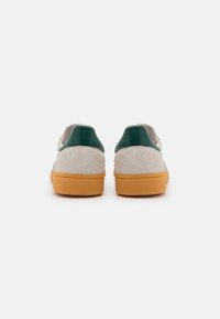 Sneakers featuring a beige suede upper, green leather accents on the heel, and a textured orange rubber sole. Rear view.