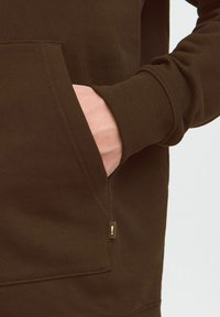 Brun sweatshirt lavet af blødt stof, med en frontlomme og ribbede manchetter. Bemærkelsesværdig detalje inkluderer et lille guldmærke på siden.
