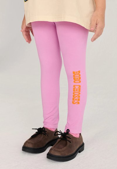 Enfant portant des leggings roses avec un texte orange "BOBO CHOSES" sur une jambe et des chaussures marron à lacets sur fond clair.