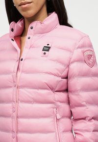 Γυναίκα που φοράει ανοιχτό ροζ μπουφάν με επένδυση, κουμπιά τύπου snap, τσέπη με φερμουάρ και ετικέτες με το λογότυπο "Blauer U.S.A." στο στήθος και το  μανίκι.