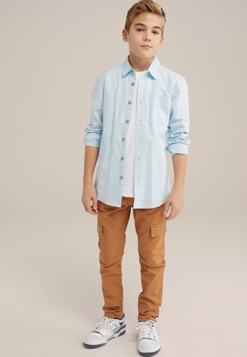 Světle modrá košile na knoflíky s kapsou, spárovaná s hnědými cargo kalhotami s bočními kapsami a bílými teniskami s modrými akcenty.